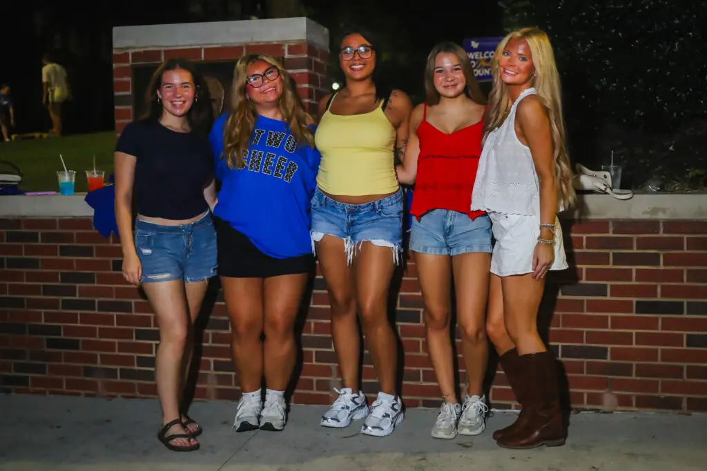 Group of five young women posing together outdoors at night in casual summer clothing in front of a brick wall.
