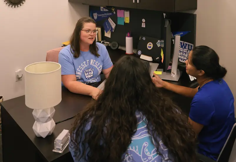Advisor sits with two students at a university office desk, discussing at the counter with badges and campus decor in view