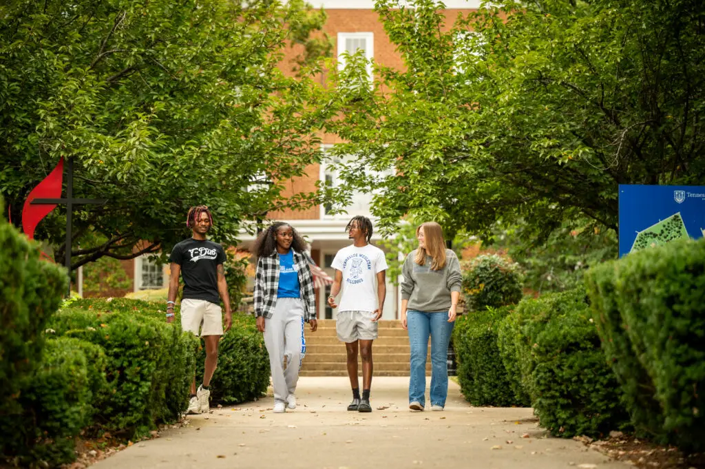 Five students walk down a campus path framed by lush greenery, brick buildings in the background.