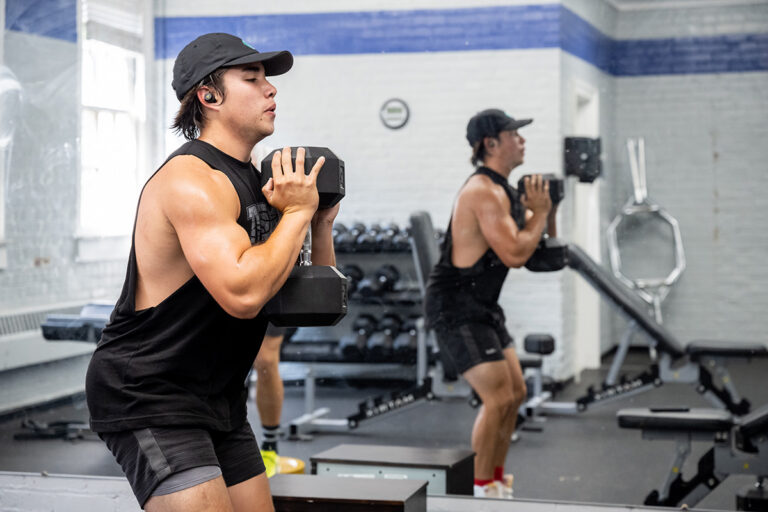 Man wearing a black tank top and cap performs a dumbbell exercise in a gym, facing a mirror.