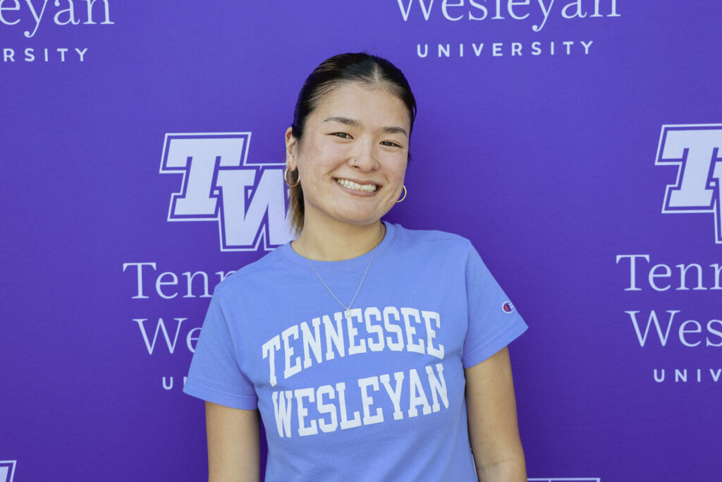 Smiling woman wearing a Tennessee Wesleyan t‑shirt stands in front of a purple university backdrop.
