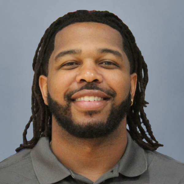 Smiling man with shoulder-length dreadlocks wearing a gray polo against a blue background.