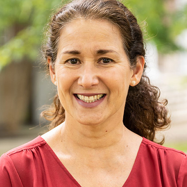Smiling woman with dark curly hair wearing a red top, outdoors with greenery in the background.