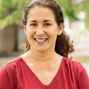 Smiling woman with brown hair tied back, wearing a red blouse, standing outdoors on a sunny day.