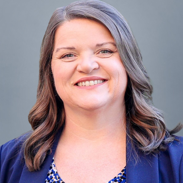 Professional head-and-shoulders portrait of a smiling woman with shoulder-length gray-brown hair, wearing a blue blazer against a neutral backdrop.
