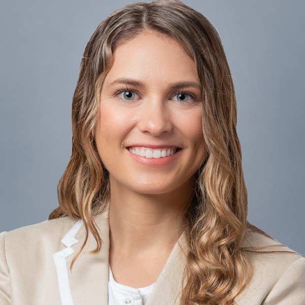 Portrait of a smiling woman with wavy light brown hair, wearing a beige blazer against a gray backdrop