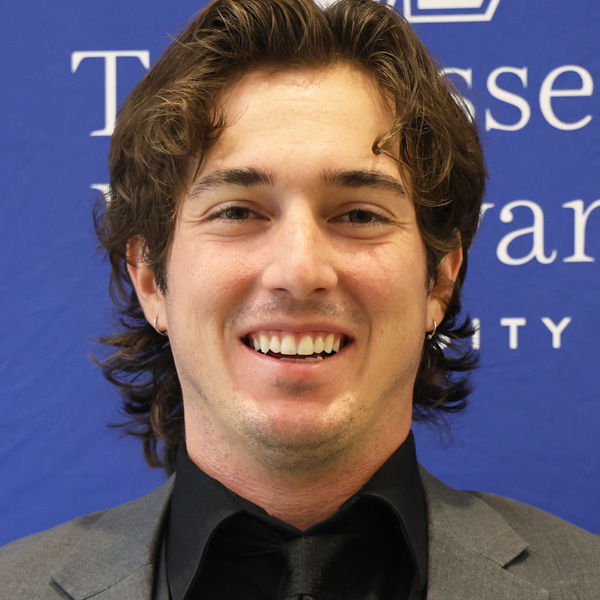 Smiling man in a gray suit and black shirt, standing in front of a blue backdrop with white text.