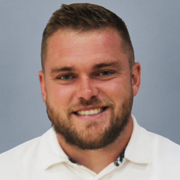 Close-up portrait of a smiling man with short hair and a trimmed beard, wearing a white polo shirt against a gray background.