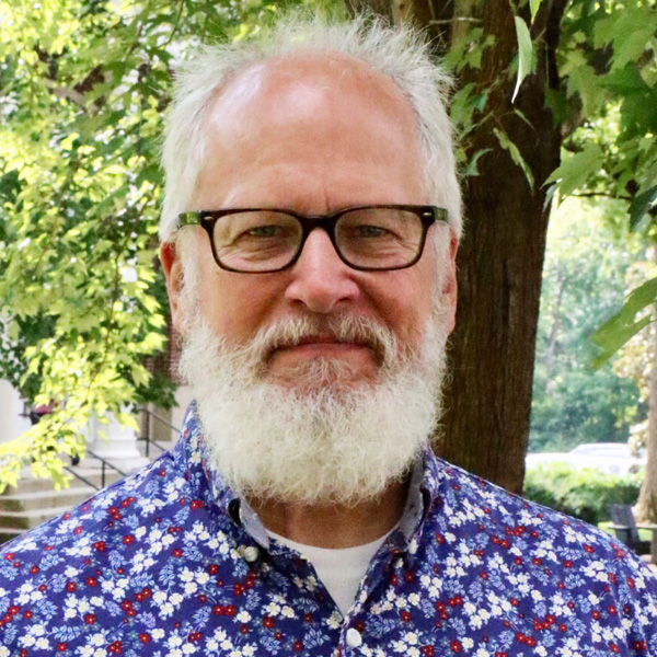 Older man with white beard and glasses smiling outdoors among trees, wearing a blue floral shirt.