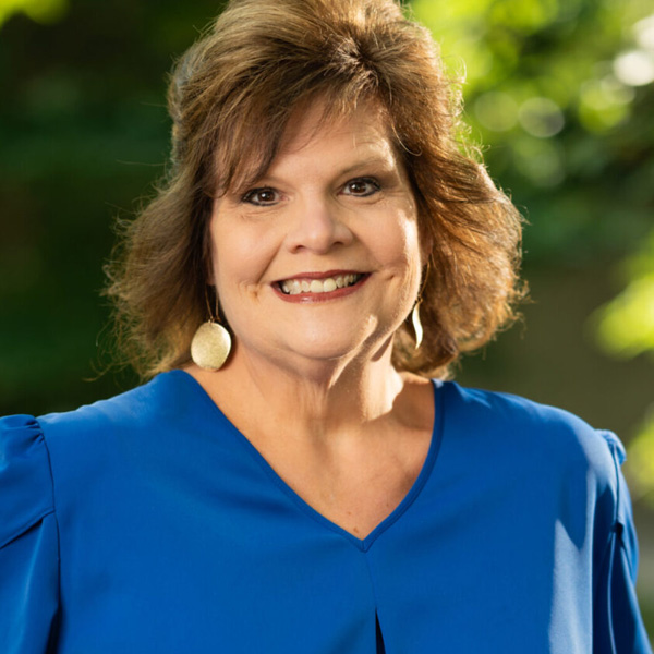 Smiling woman with short brown hair wearing a blue blouse, outdoors with greenery in the background.