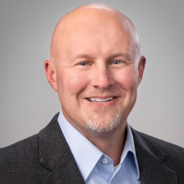 Smiling bald man wearing a dark blazer and light blue shirt, portrait against a gray background (professional headshot).
