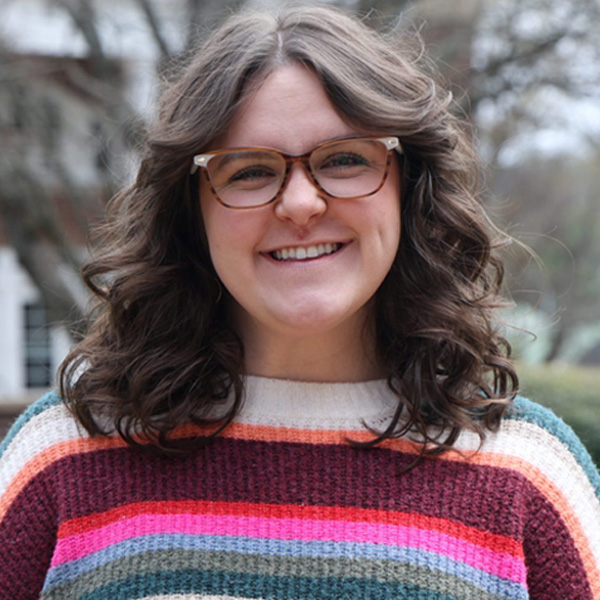 Smiling woman with wavy brown hair and brown glasses, wearing a multicolored striped sweater outdoors.
