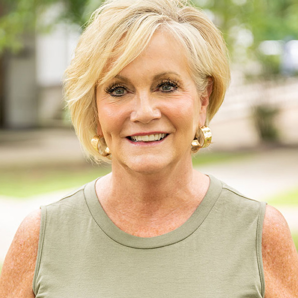 Smiling blonde woman in a green sleeveless top outdoors, gold hoop earrings, sunny day.