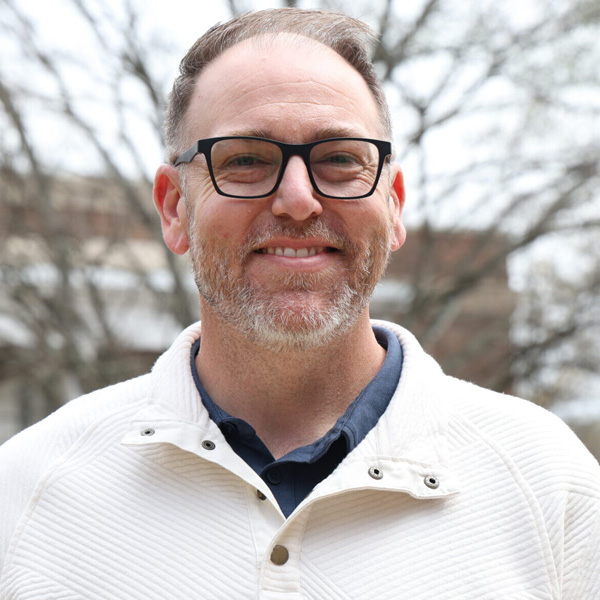 Smiling man with short gray beard and black glasses outdoors on a cloudy day.