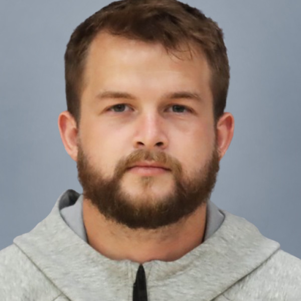 Portrait of a man with short brown hair and a full beard, wearing a light gray hoodie against a gray background, looking at the camera with a neutral expression.
