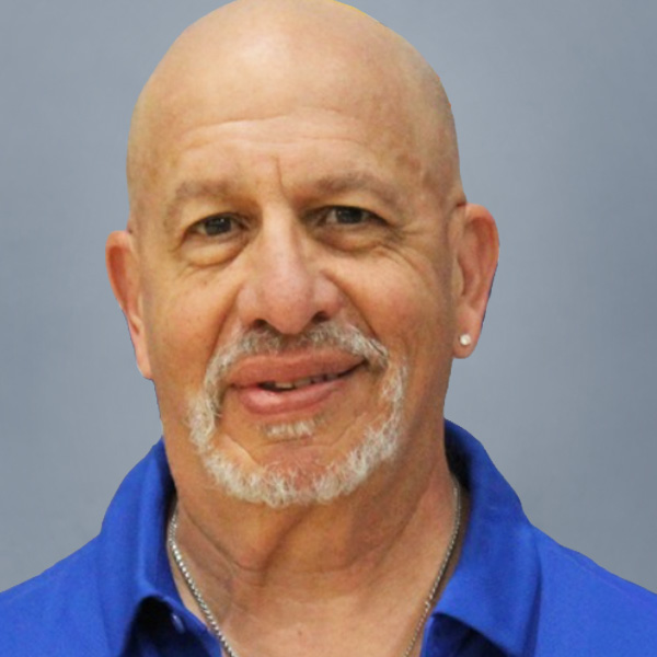 Smiling bald man with a white goatee wearing a blue collared shirt against a gray background.