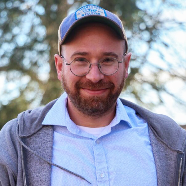 Smiling man with glasses and a beard wearing a blue cap with a Wesleyan logo, a light blue shirt, and a gray hoodie outdoors.