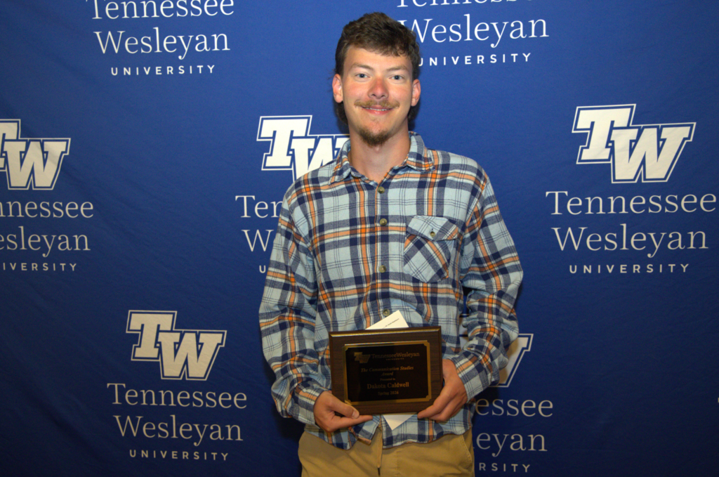 Young man in a blue plaid shirt holds a Tennessee Wesleyan University award plaque at a campus event, smiling for the photo.