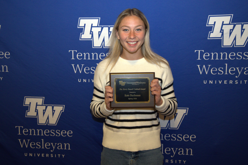 Young woman smiling and holding a framed award plaque in front of a Tennessee Wesleyan University backdrop.