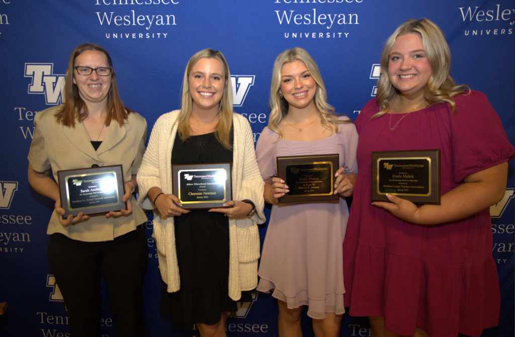 Four smiling young women stand in front of a Tennessee Wesleyan backdrop, each holding a plaque of recognition.