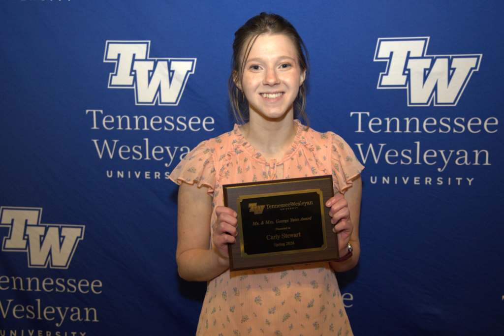 Smiling student Carly Stewart holds a Tennessee Wesleyan University award plaque in front of a blue TWU backdrop.