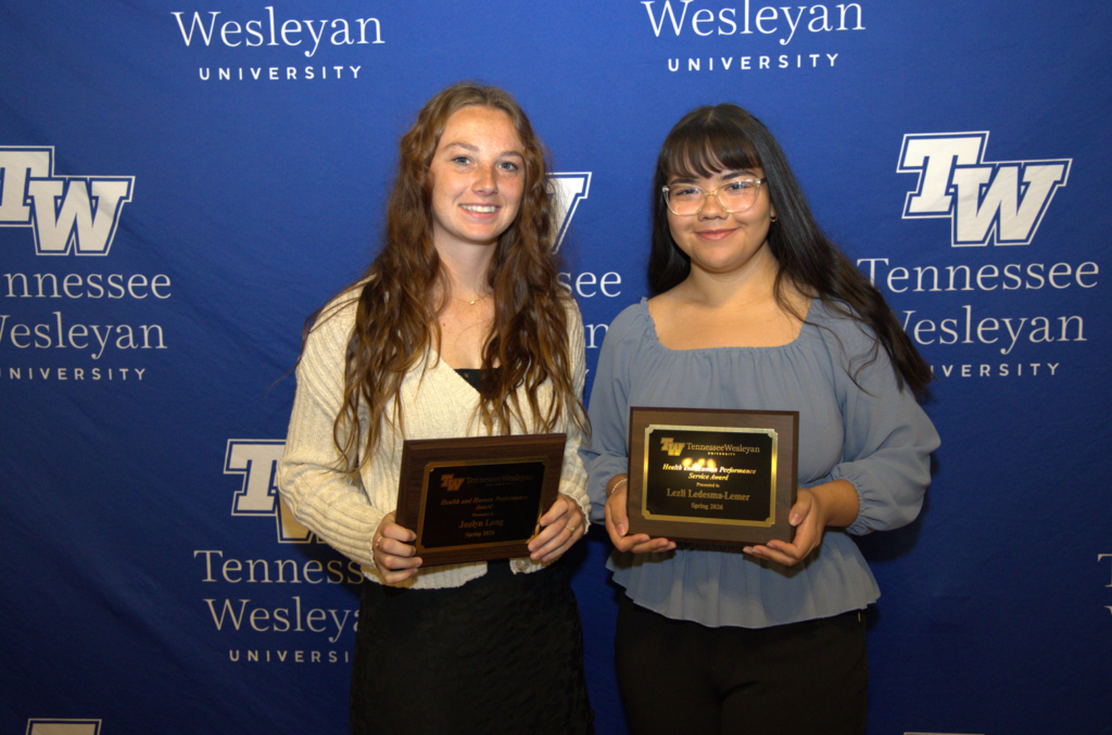 Two young women stand in front of a blue Tennessee Wesleyan backdrop, each holding a wooden plaque award.
