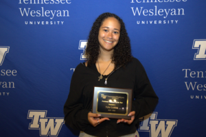 Smiling young woman with curly hair holding a rectangular award plaque in front of a Tennessee Wesleyan University backdrop.