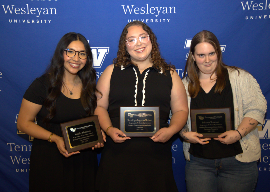Three female students stand together holding plaques at a Wesleyan University awards event, smiling in front of a blue backdrop with university logos.