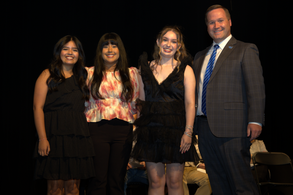 Four people stand on a stage, smiling for a photo: three women in dresses and a man in a suit beside them.