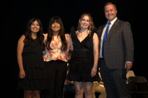 Four people stand on a stage, smiling for a photo: three women in dresses and a man in a suit beside them.