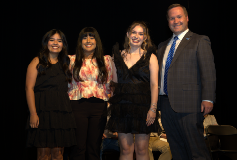 Four people stand on a stage, smiling for a photo: three women in dresses and a man in a suit beside them.