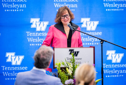 Speaker at a podium in a pink blazer speaking to an audience, with a blue Tennessee Wesleyan University backdrop and microphone in front of her.