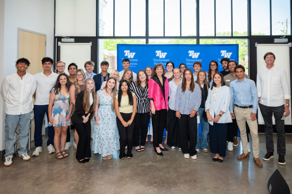 Group of diverse students posing with a woman in a bright pink blazer in front of a Tennessee Wesleyan University banner during a campus event.