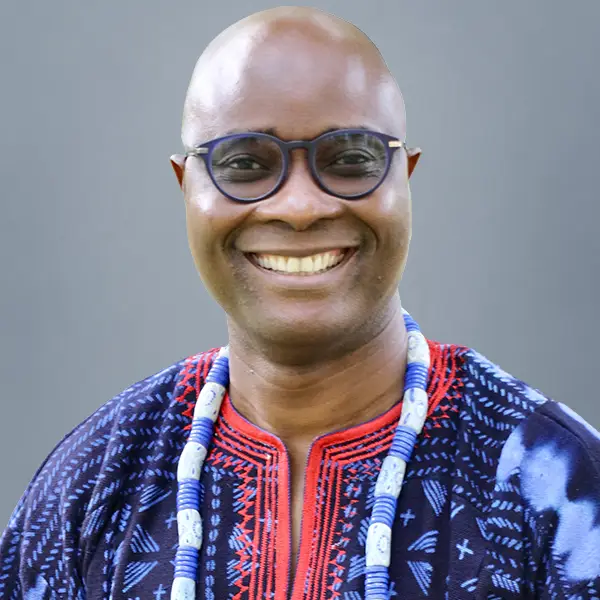 Smiling man wearing round glasses and a blue patterned traditional shirt with a beaded necklace.
