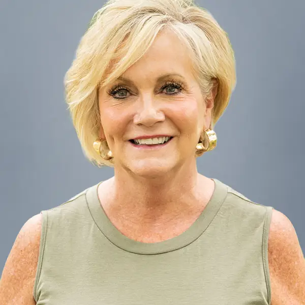 Smiling older woman with short blonde hair wearing a sage green sleeveless top and gold hoop earrings, against a gray-blue background.