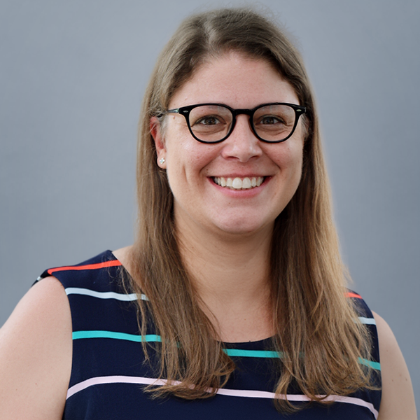Smiling woman with shoulder-length brown hair and black glasses, wearing a navy striped sleeveless top, against a gray backdrop.