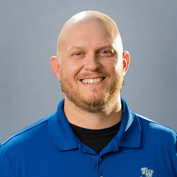 Smiling bald man with a light beard wearing a blue polo shirt against a gray background.