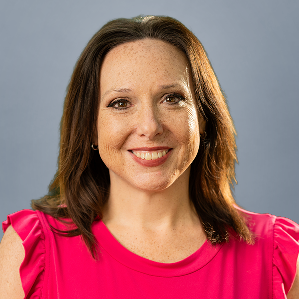 Portrait of a smiling woman with shoulder-length brown hair in a pink blouse against a blue backdrop, facing the camera.