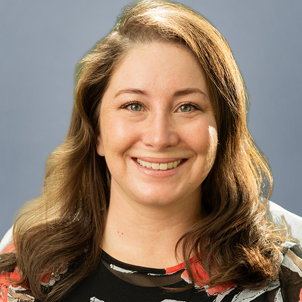 Portrait of a smiling woman with light brown hair wearing a floral top, against a blue-gray background.