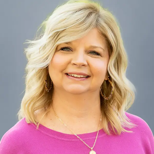 Smiling blonde woman in a pink sweater with hoop earrings, facing the camera against a gray background.