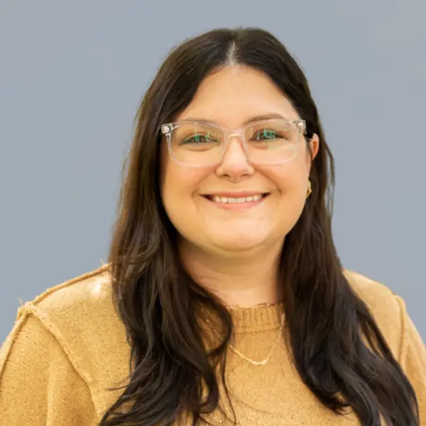 Woman with long dark hair and clear glasses smiling at the camera, wearing a tan sweater against a gray background, portrait.