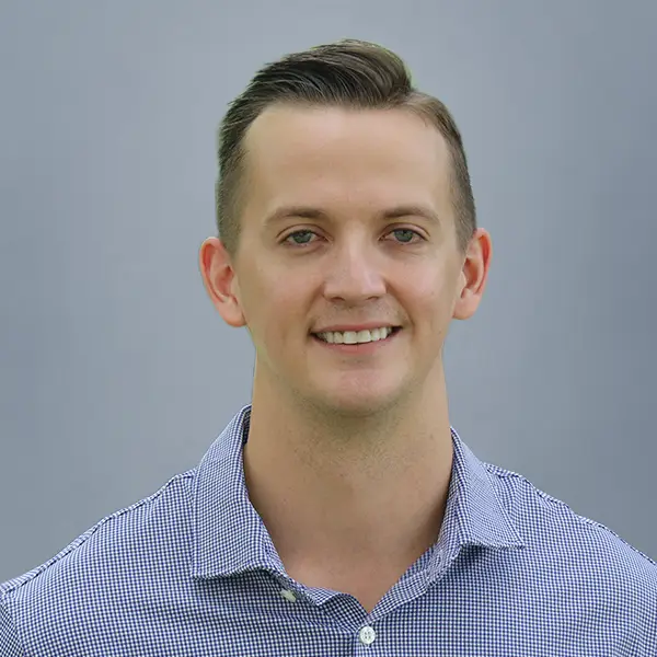 Head-and-shoulders portrait of a smiling man in a blue checkered shirt against a gray backdrop.