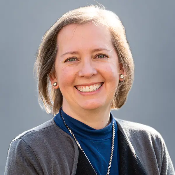 Smiling woman with short light brown hair wearing a blue turtleneck and gray cardigan, neutral gray backdrop.
