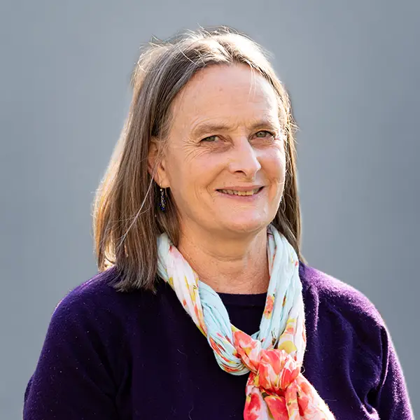 Smiling woman with shoulder-length gray-brown hair wearing a purple sweater and a floral scarf, posed against a gray background.