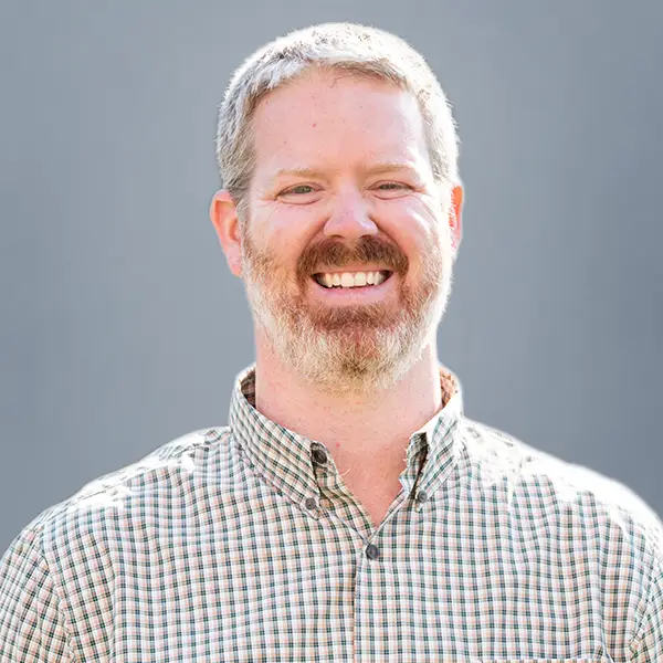 Smiling man with short gray hair and beard wearing a checkered button-down shirt against a gray background.