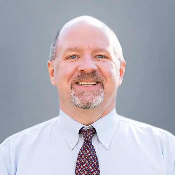Smiling middle-aged man with a light beard in a light blue shirt and patterned tie against a gray background, professional headshot.