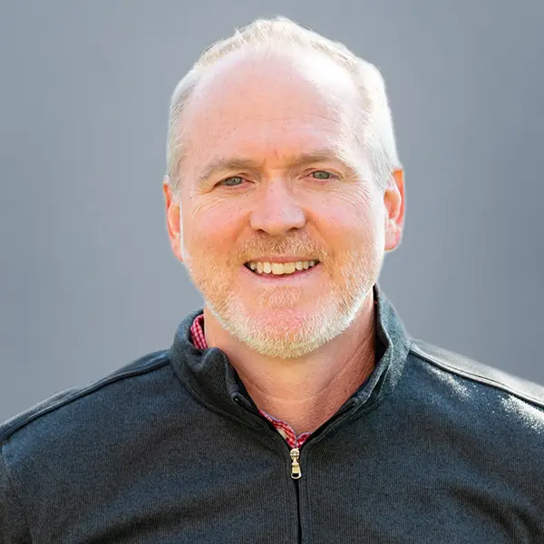 Smiling man with short white hair and a light beard, wearing a dark zip-up jacket, facing the camera against a gray background.