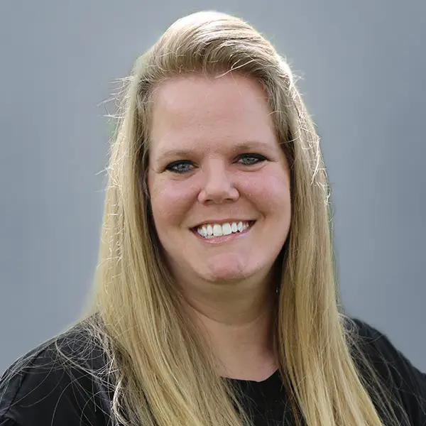 Portrait of a smiling woman with long blonde hair wearing a black top against a gray background.