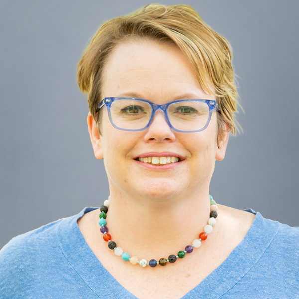 Smiling woman with short blond hair wearing blue glasses and a multicolored beaded necklace, against a gray background.