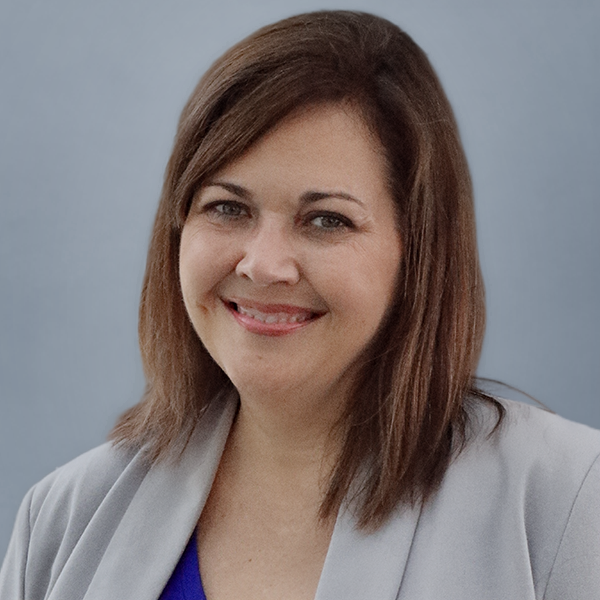 Professional headshot of a smiling woman with shoulder-length brown hair, wearing a gray blazer and blue top against a light blue backdrop.